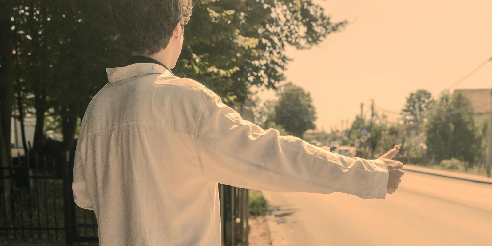 Ömer from behind, arm out and thumb up, hitchhiking on a tree-lined road