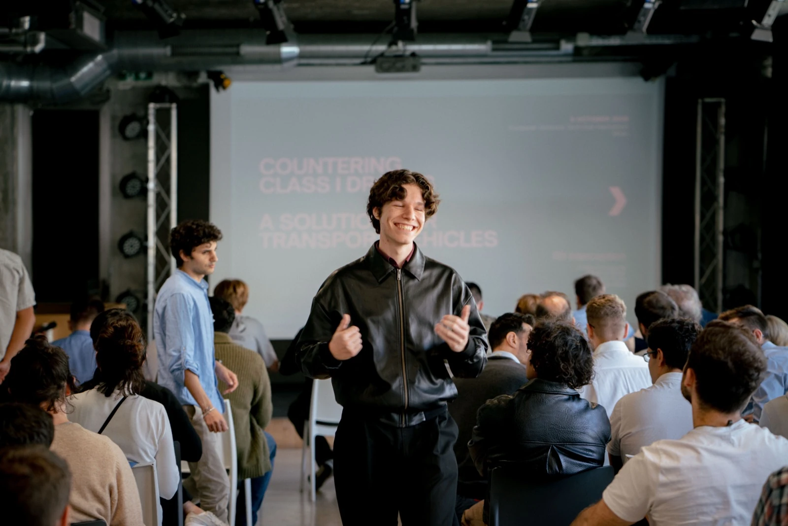 Ömer standing among a seated audience with both thumbs up, in front of a presentation screen