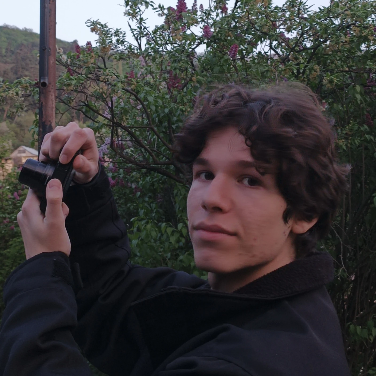 Ömer raising a compact camera to frame a shot, lilac in bloom and hills behind him at dusk